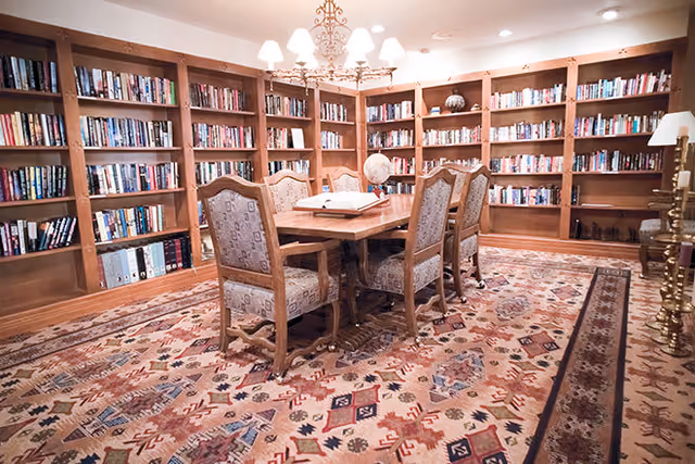 A cozy library-style room with bookshelves along the walls, a wooden table surrounded by upholstered chairs, a globe, and a patterned area rug under a chandelier.
