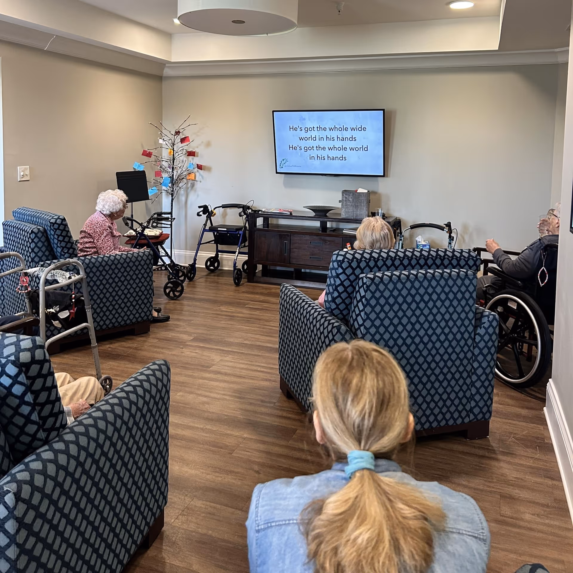 A group of elderly people sitting in a common room with patterned armchairs, watching a television mounted on the wall displaying song lyrics. Several walkers and a wheelchair are visible, and a decorative tree with colorful cards is placed near the wall.