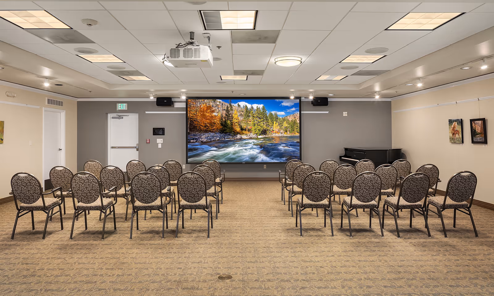 A conference or presentation room with rows of patterned chairs facing a large screen displaying a scenic image of a river flowing through a forest with autumn-colored trees and mountains in the background. The room has beige walls, a carpeted floor, ceiling lights, a piano in the corner, and framed pictures on the walls.