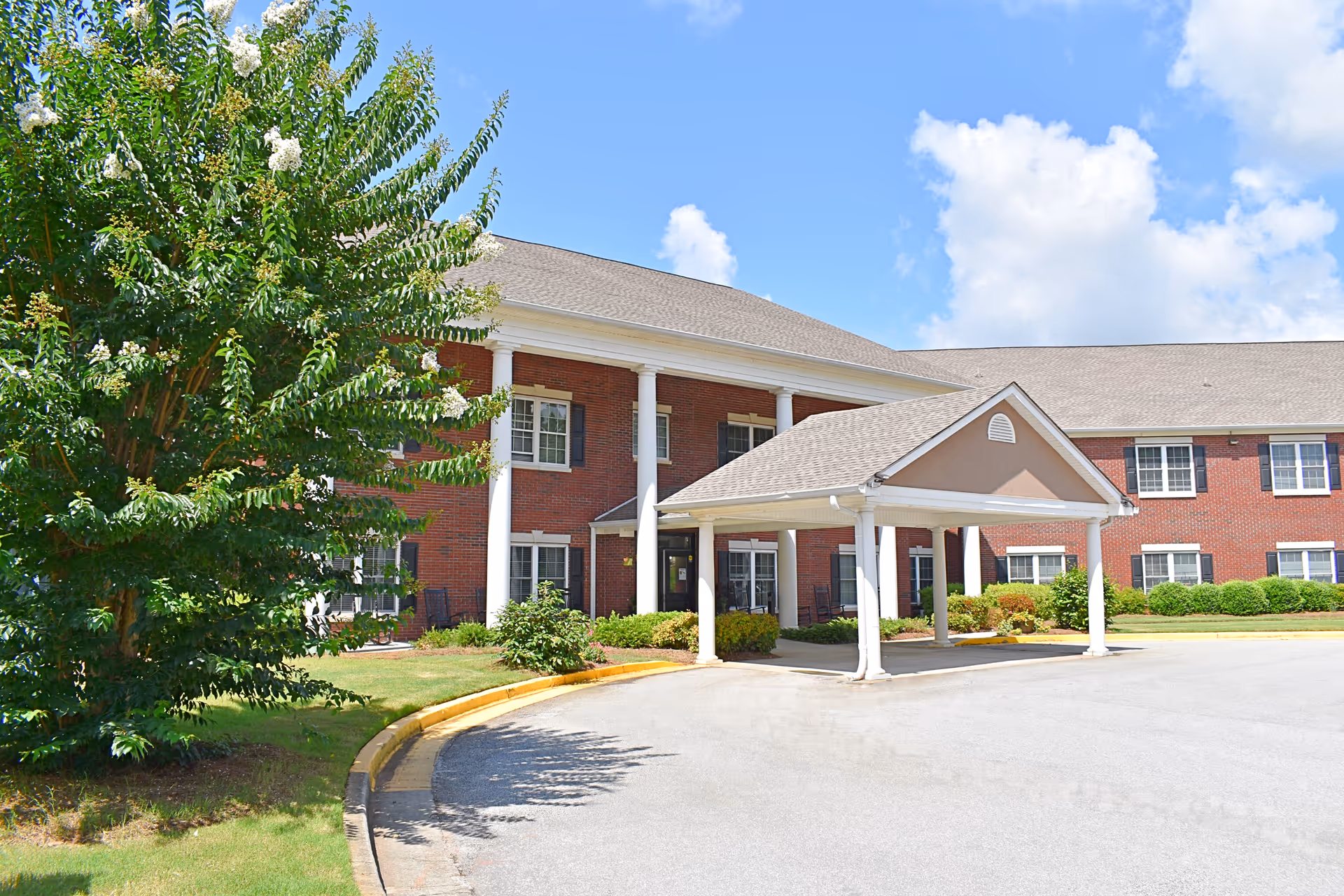 Exterior view of a two-story brick building with white columns and a covered entrance driveway under a blue sky with some clouds. There is a large green tree and some bushes near the building.