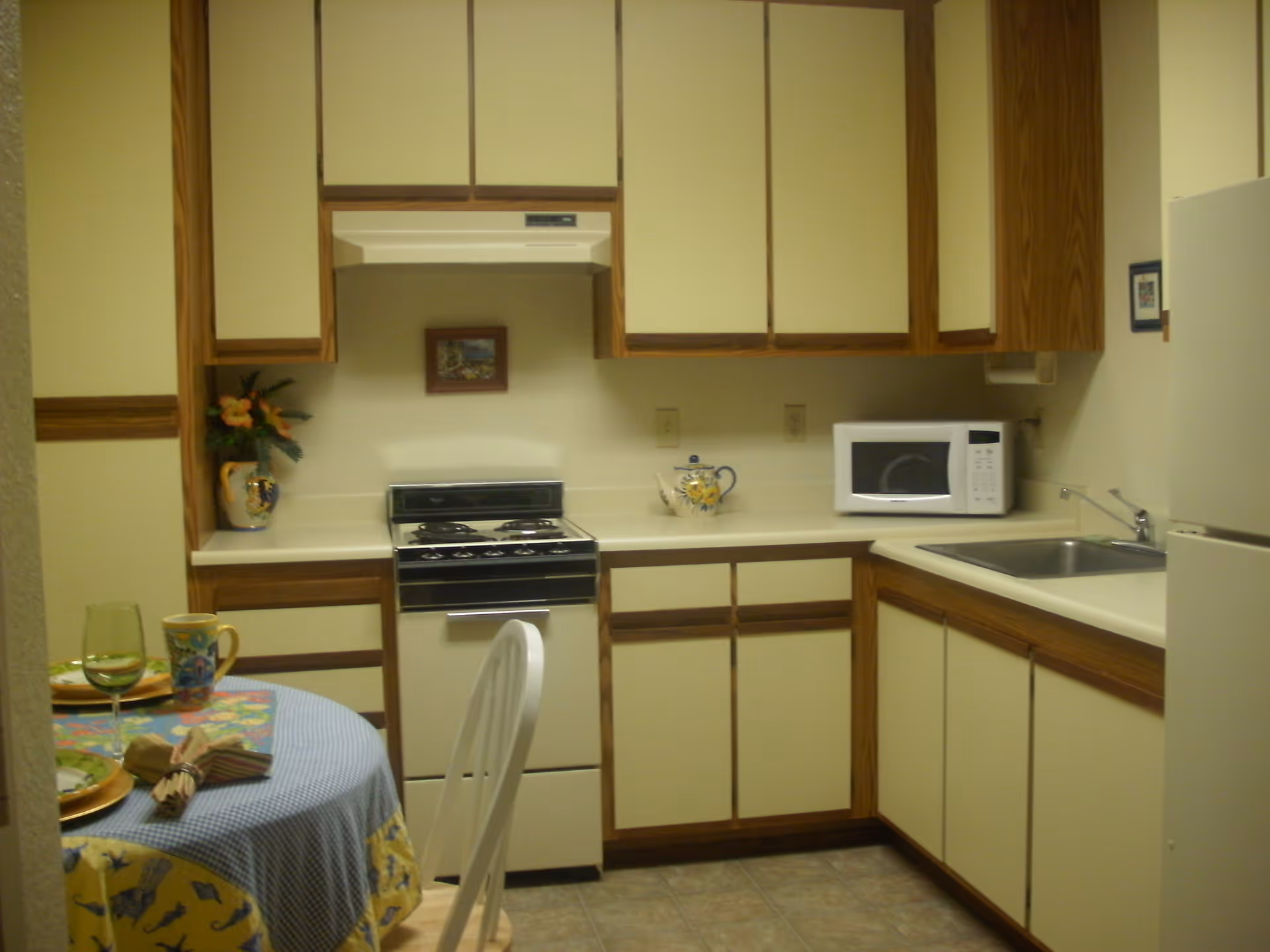 A small kitchen with beige cabinets and wood trim, a white stove with an oven, a microwave on the counter, a stainless steel sink, and a white refrigerator. A round dining table with a blue checkered tablecloth, two place settings, and a floral-patterned chair is partially visible in the foreground. There is a small framed picture on the wall above the stove and a decorative teapot on the counter.