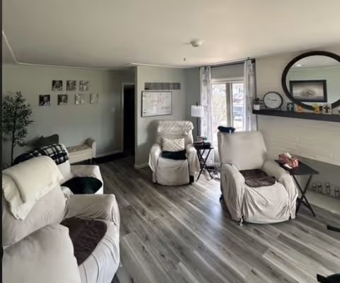 Living room with several slipcovered recliner chairs, wood‑look flooring, a fireplace mantel with a round mirror, and a window with curtains.