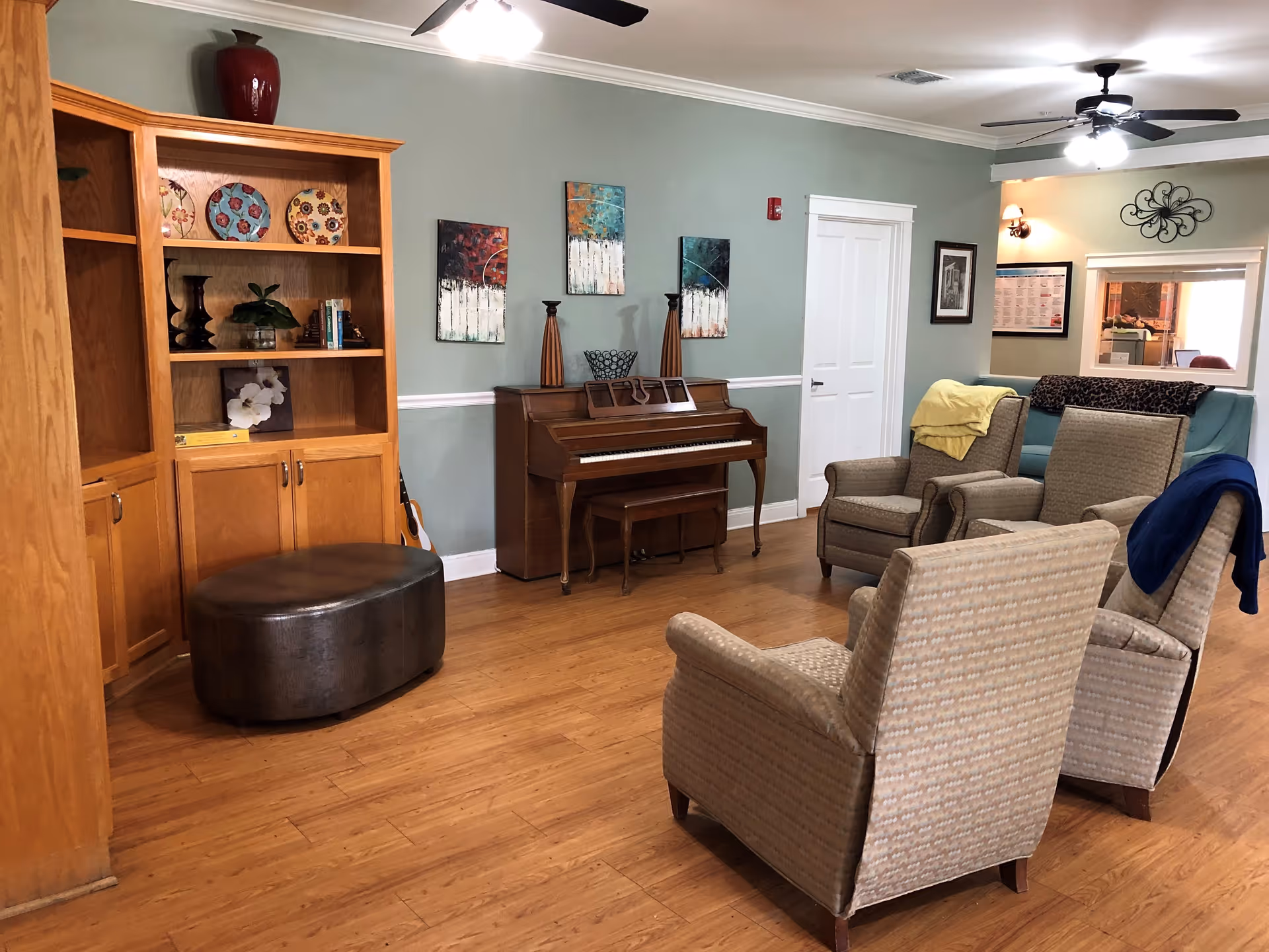 A cozy living room area with wooden flooring, light green walls, and ceiling fans. The room features a wooden piano against the wall, a wooden bookshelf with decorative plates and books, and four upholstered armchairs arranged in a semi-circle. There is a dark brown ottoman in front of the bookshelf and various wall decorations including paintings and framed pictures.
