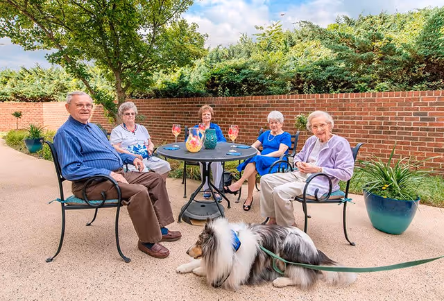 Five elderly people sitting around a round outdoor table with drinks and a pitcher, on a patio with a brick wall and greenery in the background. A large fluffy dog on a leash lies on the ground in front of the table.