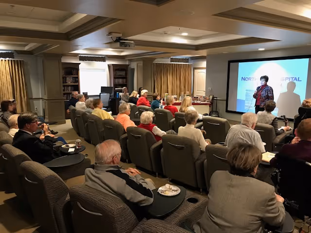 A group of elderly people seated in a room with theater-style chairs, attentively watching a presentation projected on a screen at the front. The presenter stands near the screen speaking to the audience. The room has a cozy, well-lit interior with bookshelves and curtains on the windows.
