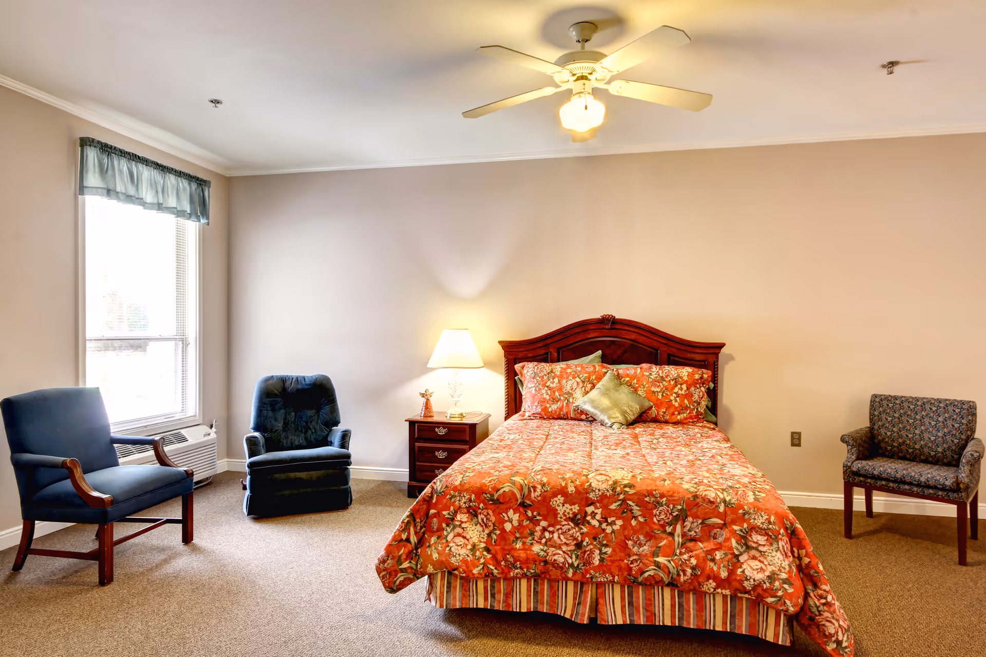 A bedroom with a floral patterned bedspread on a wooden bed frame, a nightstand with a lamp and small decorative item, three upholstered chairs, a window with a blue valance, and a ceiling fan with a light.
