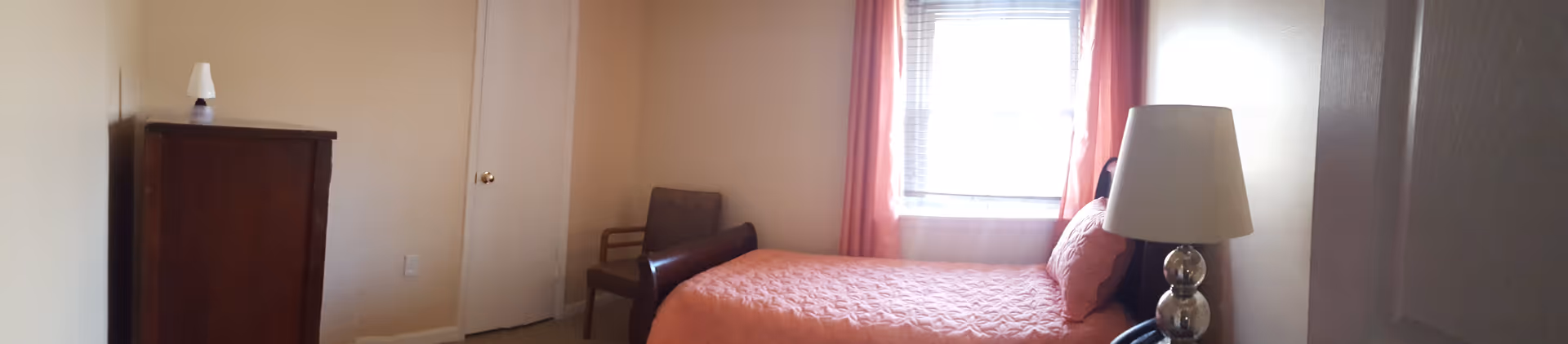 A small bedroom with a single bed covered in a coral quilt and matching pillow, a window with coral curtains letting in natural light, a wooden dresser with a small lamp on top, a brown chair, and a bedside table with a lamp.
