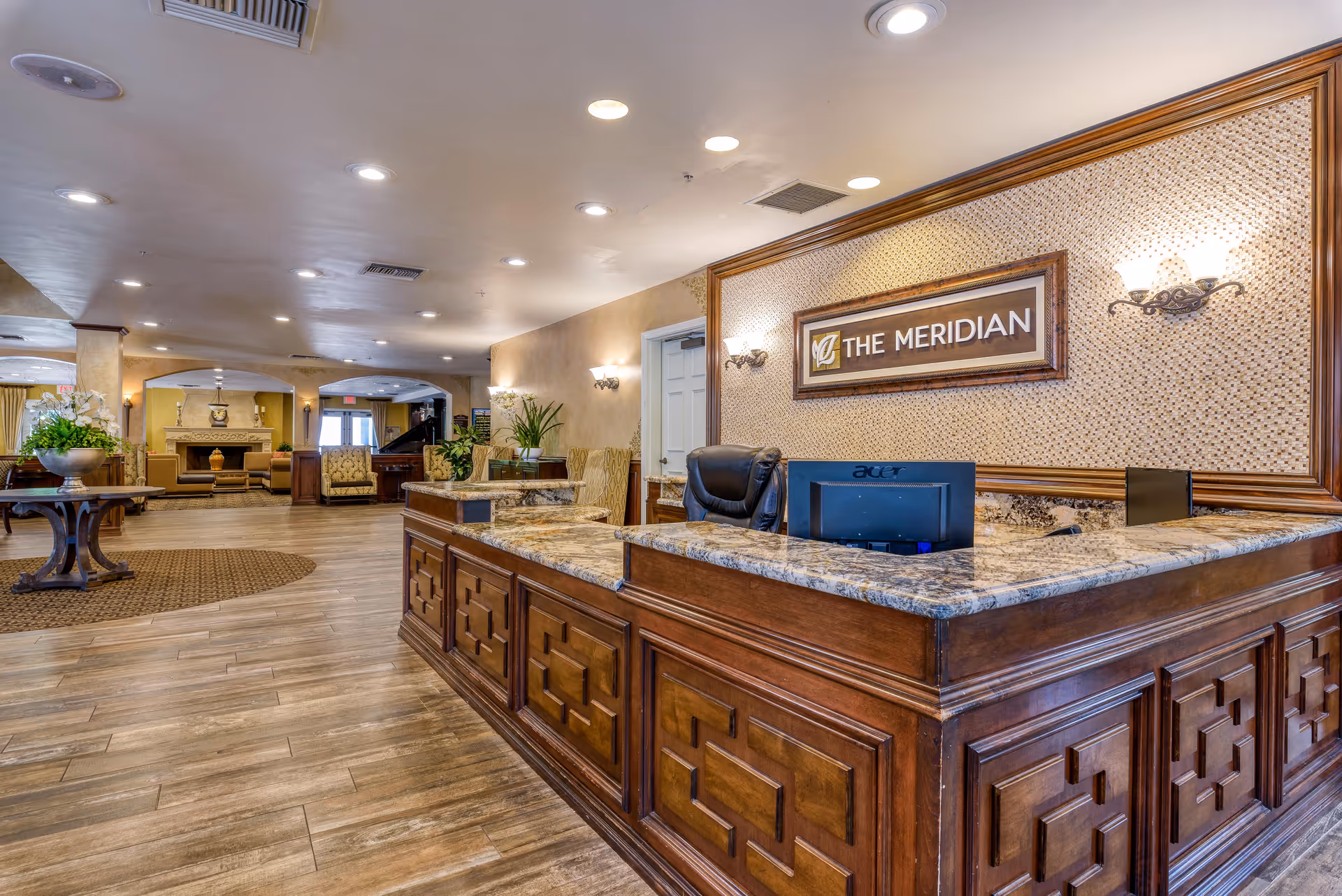 Reception area of The Meridian senior living facility with a large wooden front desk featuring a granite countertop, a computer monitor, and a black office chair. The background shows a spacious lobby with wooden flooring, a round table with a flower arrangement, comfortable seating, and warm lighting fixtures on the walls.