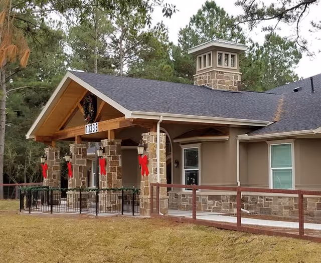 Exterior view of a single-story building with stone pillars and beige walls, decorated with red bows and a wreath, surrounded by trees and a fenced yard.