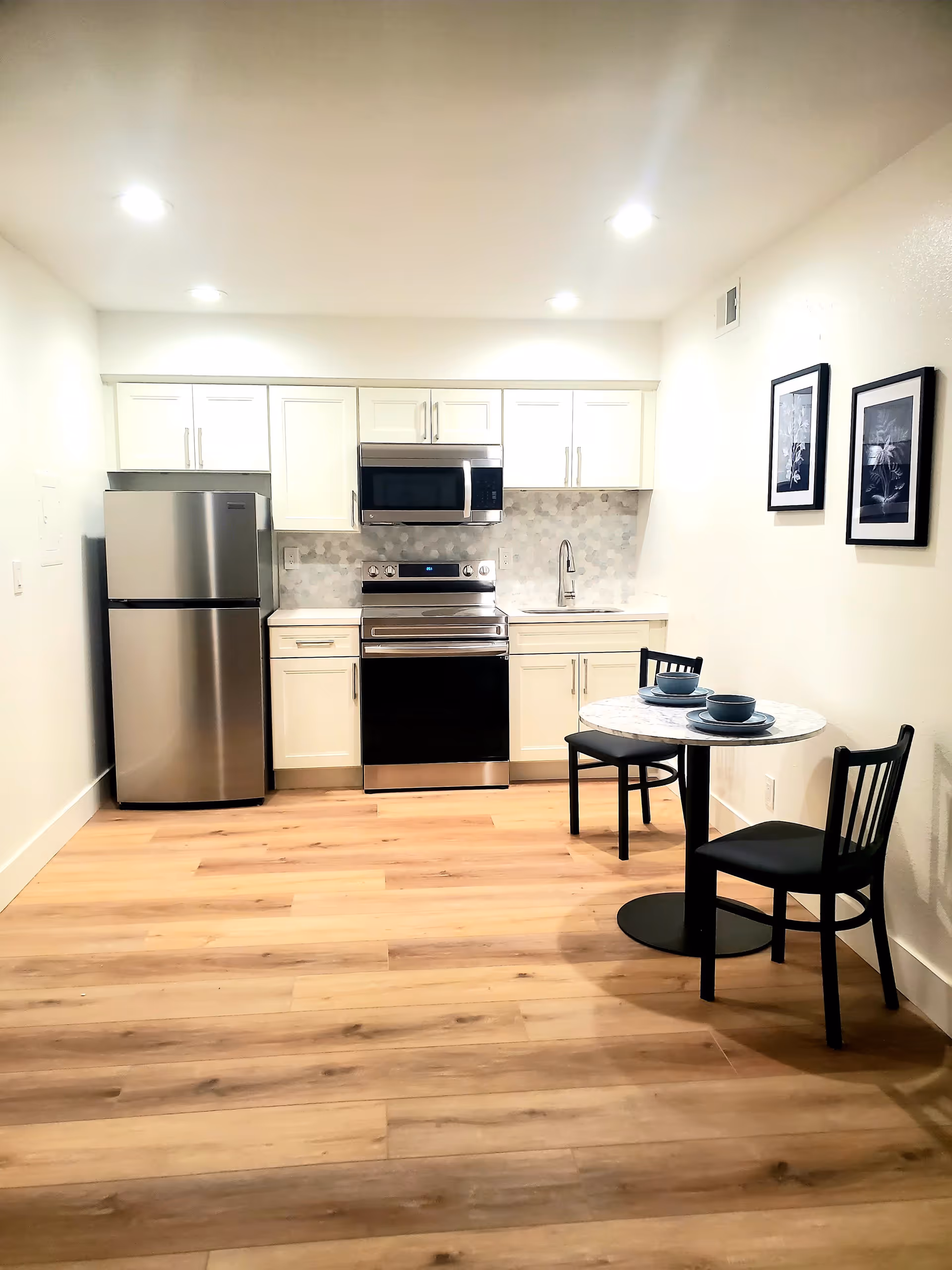 A modern kitchen area with white cabinets, a stainless steel refrigerator, stove, and microwave. There is a small round dining table with two black chairs, each set with a bowl and plate. Two framed black and white pictures hang on the wall above the dining table. The floor is light wood, and the room is well-lit with recessed ceiling lights.