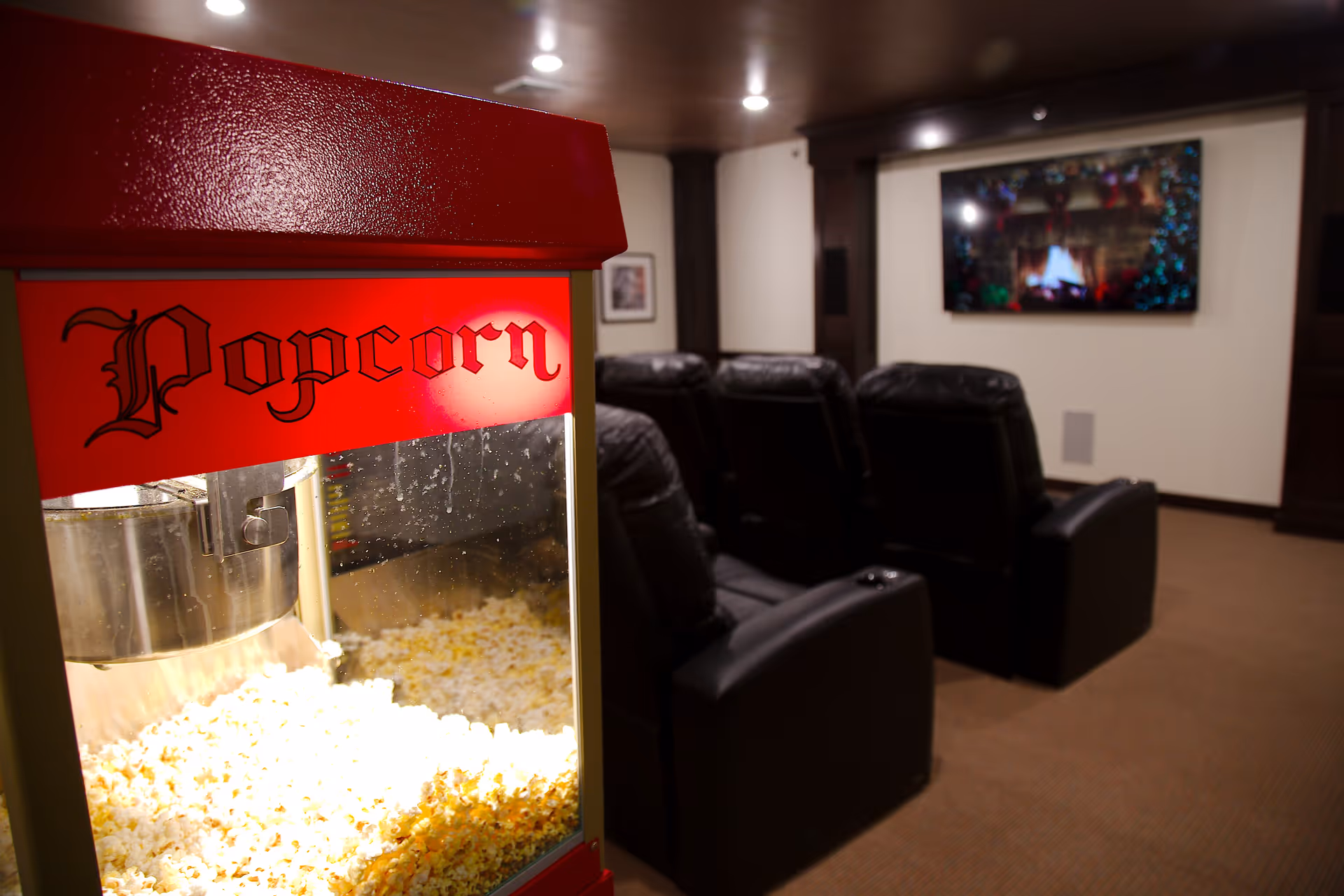 A cozy home theater room with a popcorn machine in the foreground and several black leather recliner chairs facing a large flat-screen TV mounted on the wall, which displays a festive fireplace scene.