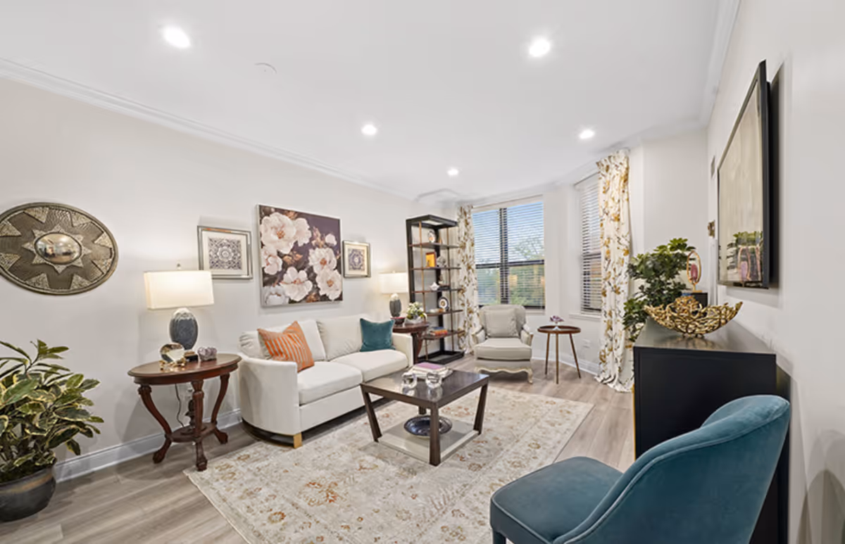 A bright and cozy living room with a white sofa adorned with orange and teal pillows, a wooden coffee table on a patterned rug, a blue armchair, and a beige armchair near windows with floral curtains. The walls are decorated with floral artwork and a decorative round wall piece. There are two side tables with lamps and plants adding greenery to the space.