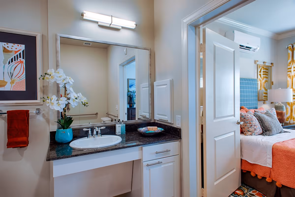 A bathroom vanity area with a large mirror, a sink with a black countertop, a blue vase with white orchids, and a red towel hanging on the wall. The bathroom opens into a bedroom with a bed featuring colorful pillows, a blue headboard, yellow patterned curtains, and a white door partially open.