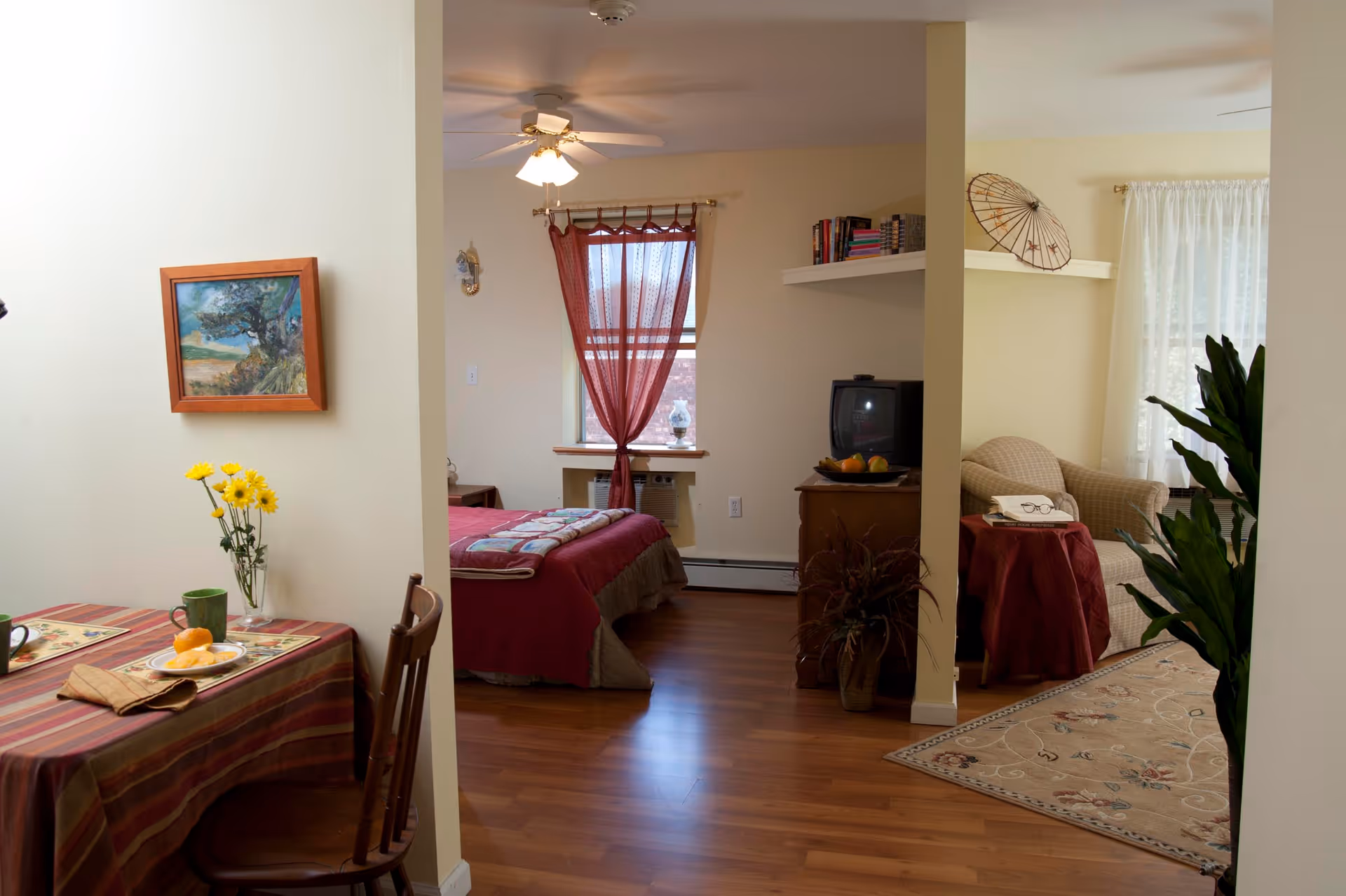 View of a cozy assisted living apartment with a small dining table set with two place settings and a vase of yellow flowers on the left. In the center, there is a bedroom with a bed covered in a red and beige quilt, a window with red curtains, and a ceiling fan. On the right, there is a living area with a beige armchair, a small table covered with a red cloth, a TV on a wooden stand, a plant, and a decorative umbrella on a shelf. The floor is wooden, and the walls are painted light yellow.