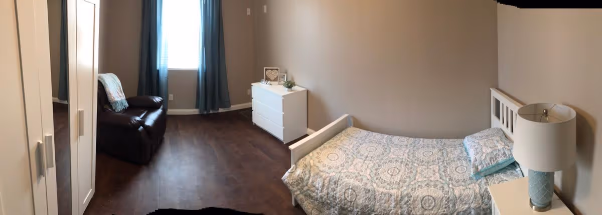 A simple bedroom with a single bed covered in a patterned quilt, a white nightstand with a lamp, a white dresser, a dark brown armchair with a blanket draped over it, and a window with blue curtains. The floor is dark wood and the walls are painted beige.