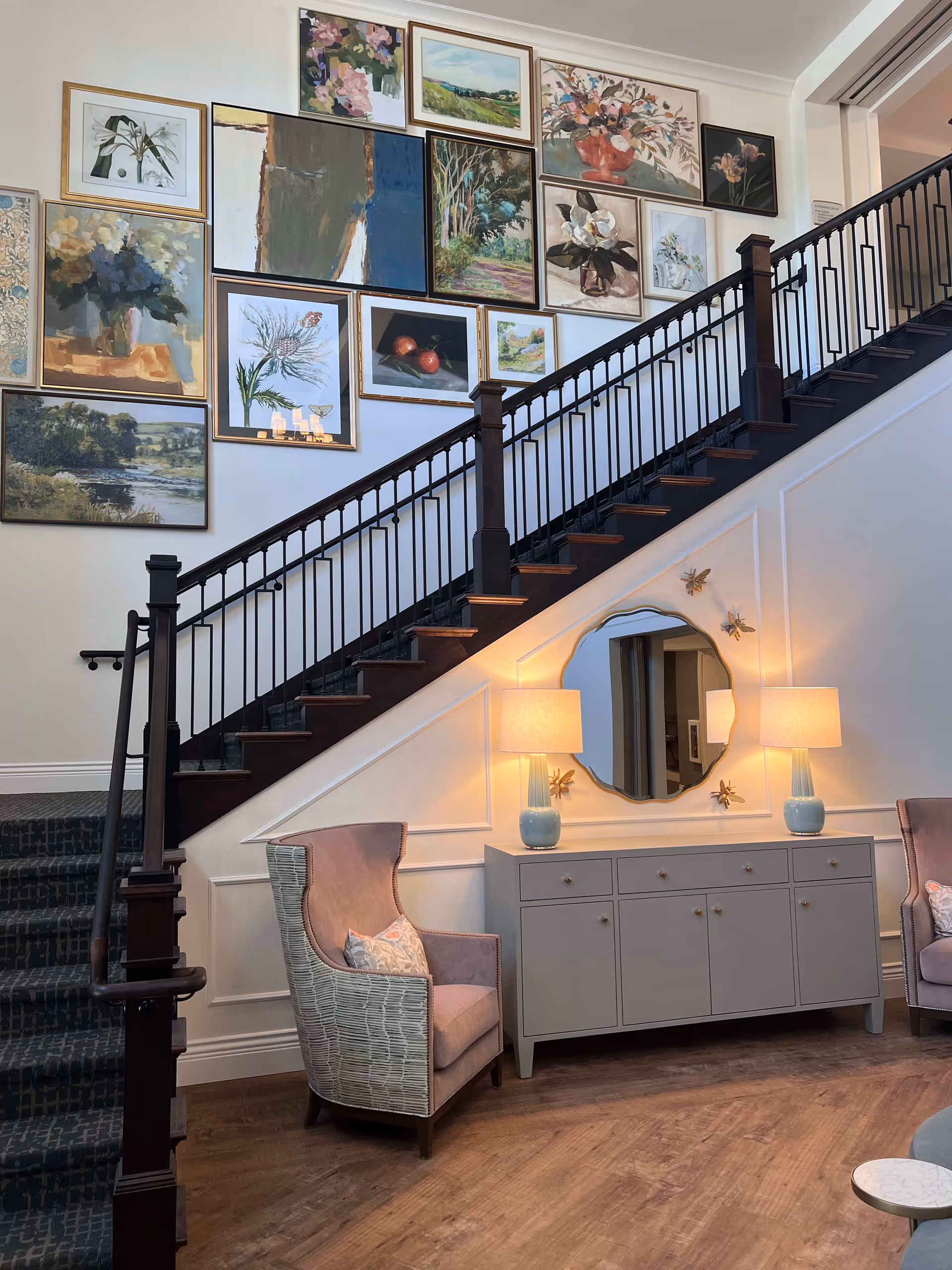 A stylish interior stairwell and seating area with a gallery wall of framed artwork, a console with lamps and mirror, and upholstered chairs.