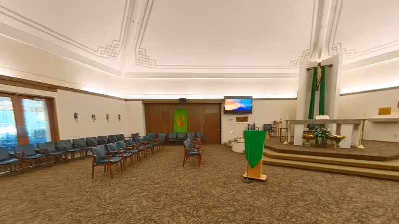 Interior view of a chapel or worship room with rows of blue chairs arranged facing a raised altar area. The altar has a crucifix and green cloth decorations. A podium with a green cloth is positioned in front of the altar. The room has a patterned carpet and a high ceiling with decorative molding. A television screen is mounted on the wall near the altar.