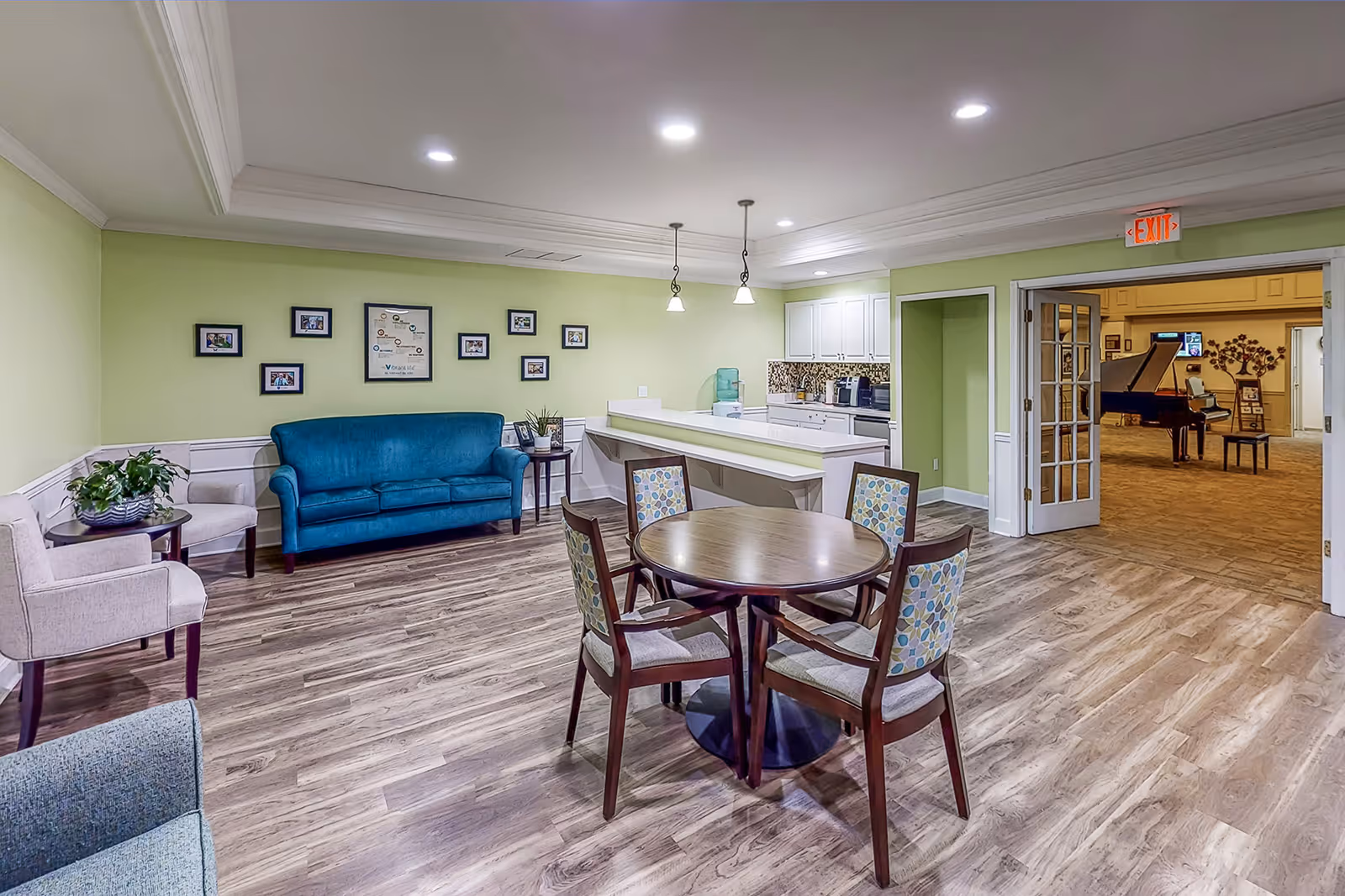 A bright and clean common area in an independent living facility featuring a round wooden table with four patterned chairs, a teal sofa, two beige armchairs, and a small kitchen area with white cabinets and a mosaic tile backsplash. The walls are painted light green, and the floor has wood-like vinyl planks. In the background, through double glass doors, a grand piano and additional seating are visible in another room.