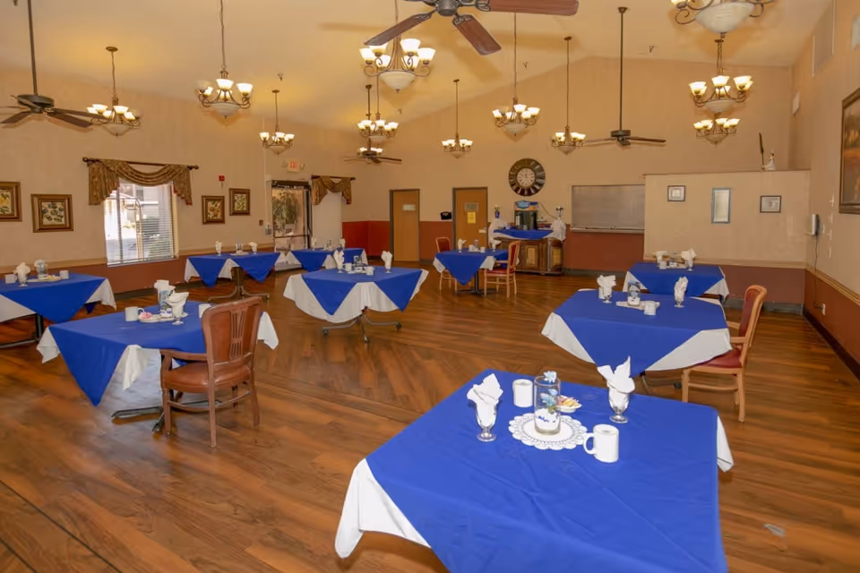 A dining room with multiple tables covered in blue and white tablecloths, each set with napkins, cups, and small centerpieces. The room has wooden floors, ceiling fans, chandeliers, framed artwork on the walls, and windows with curtains.
