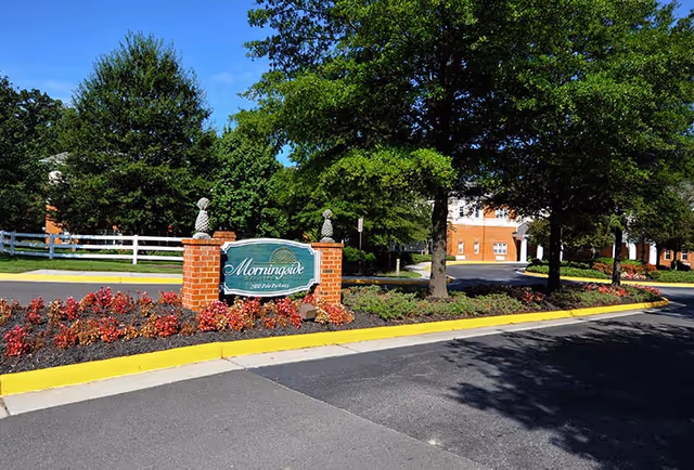 Entrance sign for Morningside of Bellgrade surrounded by landscaped flower beds and trees, with a brick building and driveway in the background under a clear blue sky.