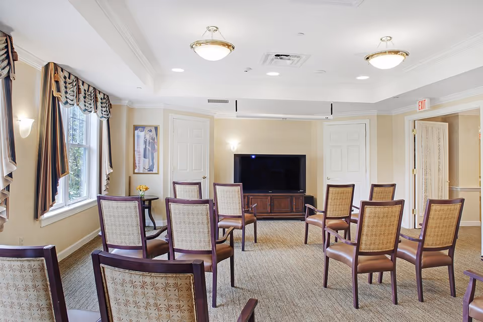 Bright common room with rows of chairs arranged facing a large TV and windows with draped curtains.