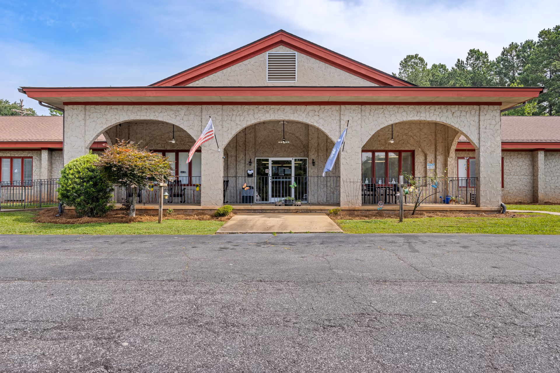 Front exterior view of a single-story building with a textured beige facade and red trim. The building has three large archways leading to a covered porch area with two flags displayed, one American and one blue flag. There are bushes and small trees in front of the building, and a paved driveway or parking area in the foreground.