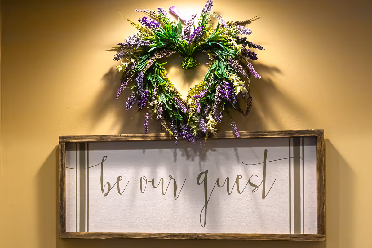 A decorative wall display featuring a heart-shaped wreath made of green leaves and purple flowers above a framed sign with the cursive text 'be our guest' on a beige wall.