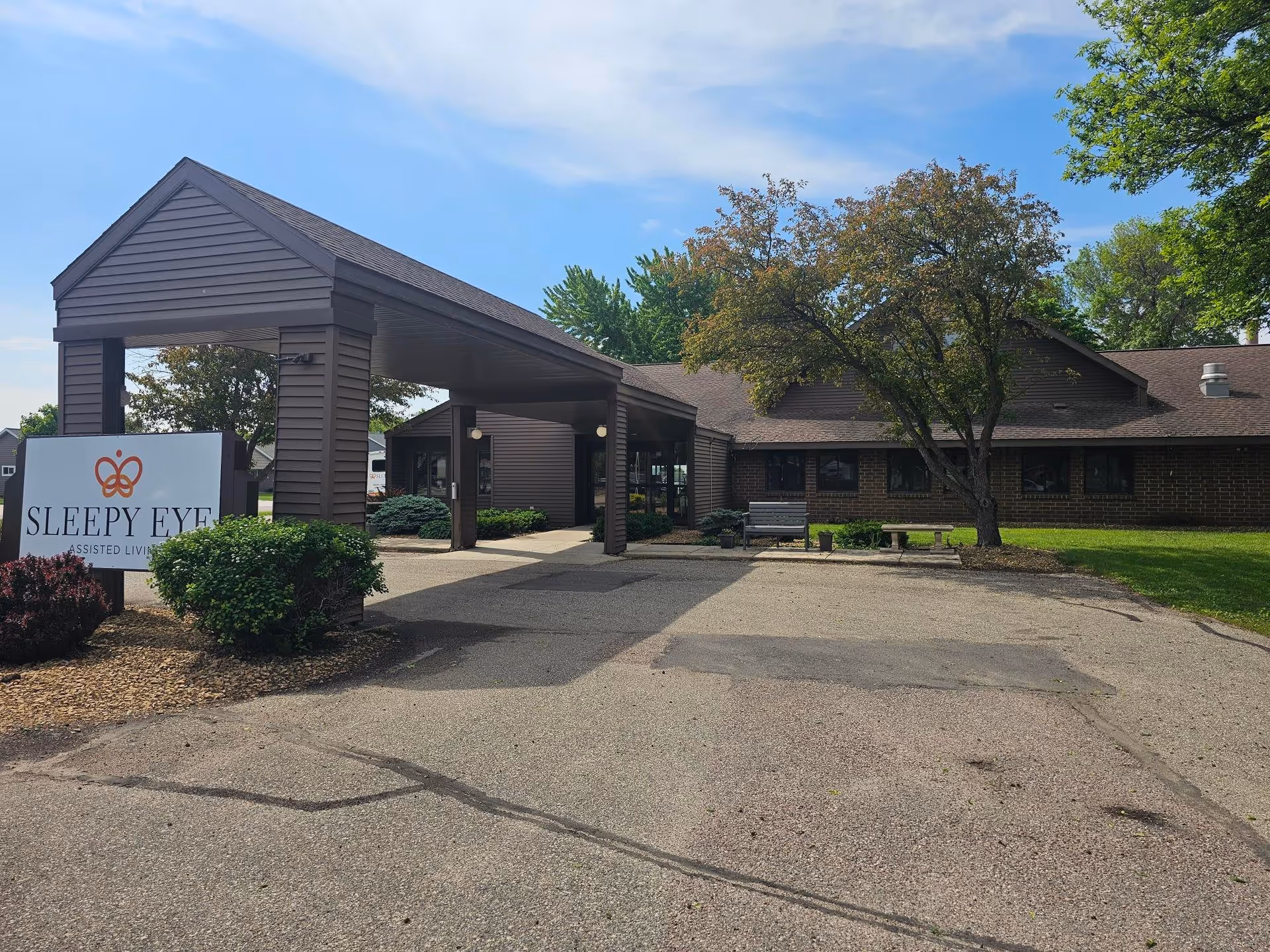 Front entrance of Sleepy Eye Assisted Living showing a covered driveway, building facade, sign, bench, and trees.