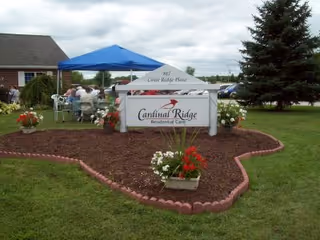 Outdoor front sign for Cardinal Ridge Residential Care with a canopy, people seated nearby, and landscaped lawn.