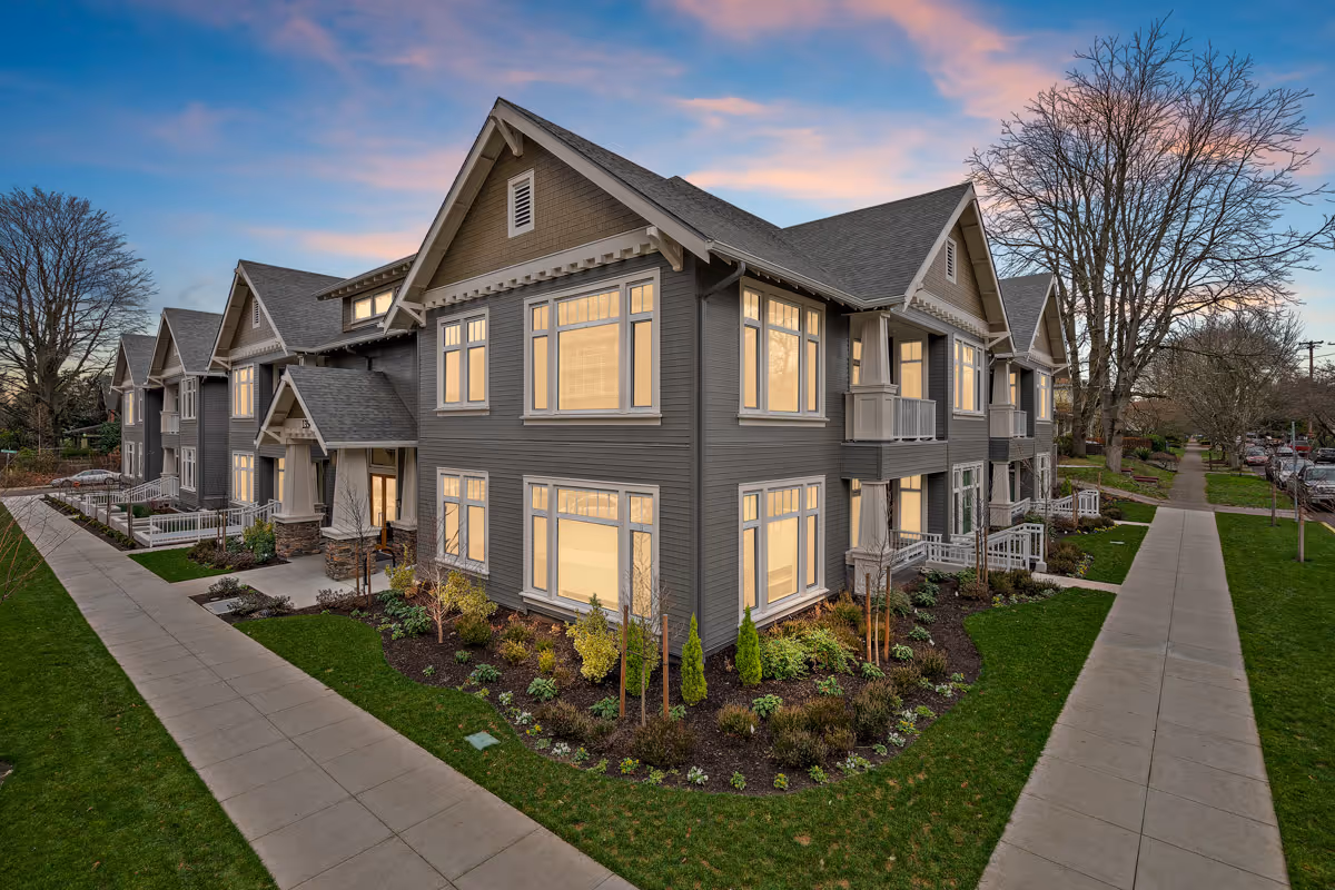 A large gray two-story residential building with lit windows and landscaped walkways at dusk.