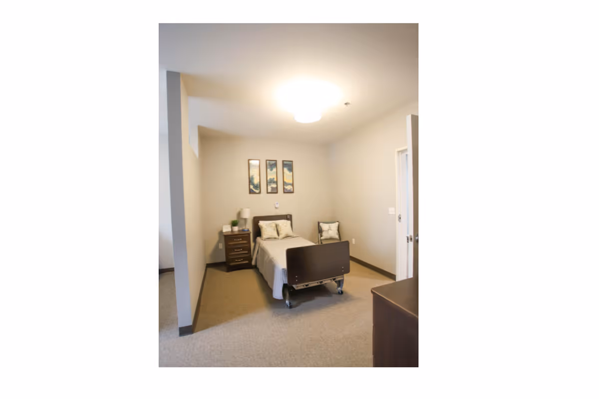 A simple hospital-style bedroom with a bed, nightstand, chairs, and neutral walls.