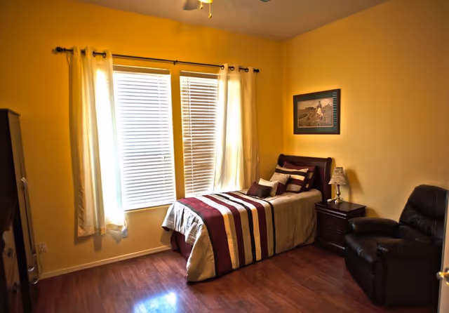 Sunlit bedroom with a single bed with striped bedding next to a nightstand and recliner, yellow walls and a window with blinds and curtains.
