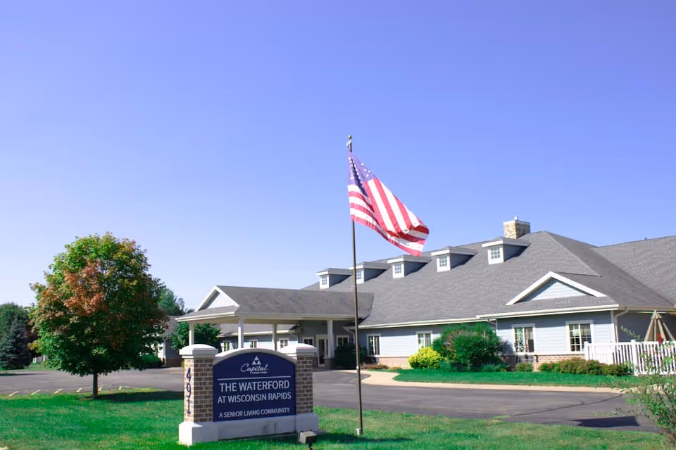 Exterior view of The Waterford At Wisconsin Rapids senior living community building with a clear blue sky, an American flag on a flagpole, a tree with green and some red leaves, and a sign in front displaying the facility's name.
