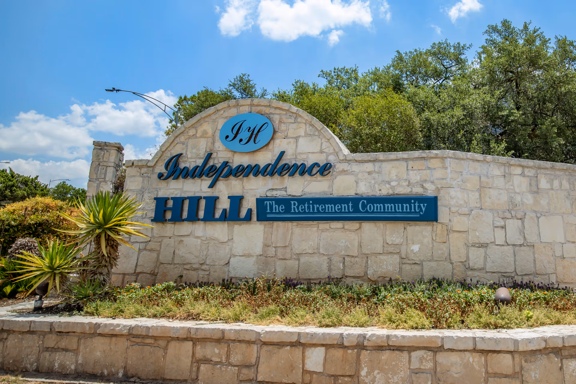 Stone entrance sign for Independence Hill Retirement Community with green plants and trees in the background under a blue sky with some clouds.