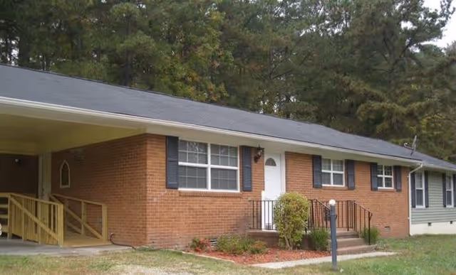 Front exterior of a single-story brick assisted living building with a white entry door, windows, small steps, a wheelchair ramp, and trees in the background.