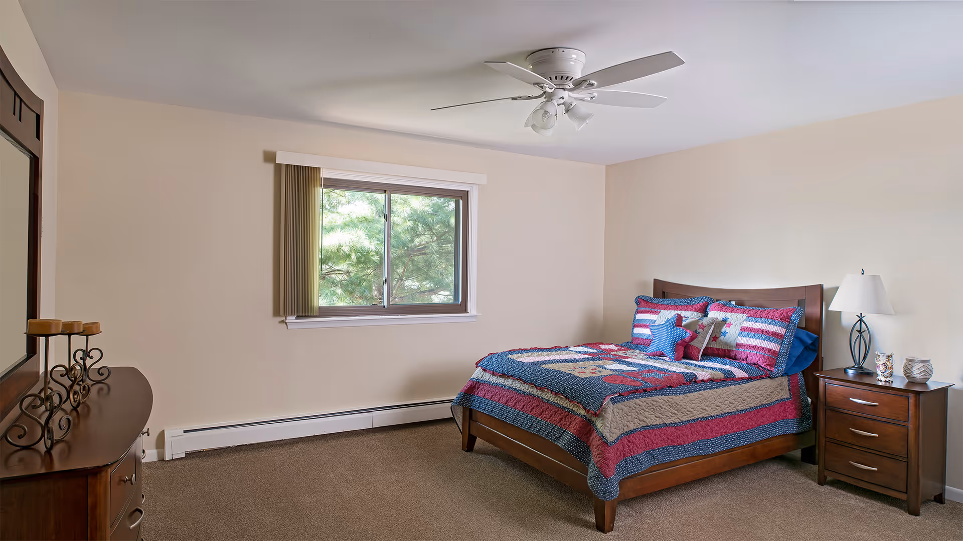 A bedroom with a wooden bed frame and a colorful quilted bedspread featuring red, blue, and beige stripes and star patterns. There are matching pillows on the bed. Next to the bed is a wooden nightstand with a white lamp and decorative items. A window with vertical blinds lets in natural light, and a ceiling fan with lights is mounted on the ceiling. A wooden dresser with a mirror and candle holders is visible on the left side of the room. The walls are painted beige and the floor is carpeted.