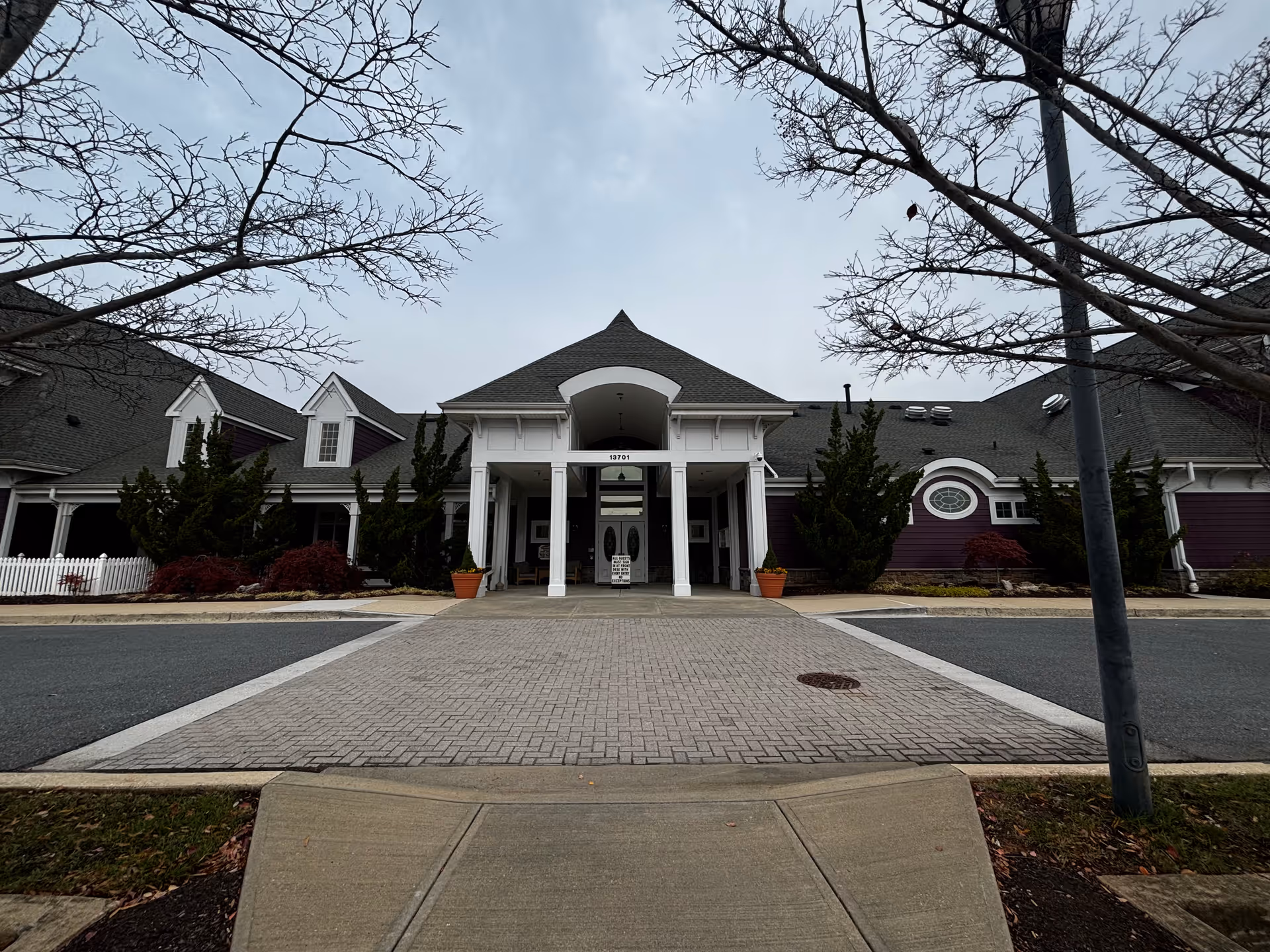 Front exterior view of a single-story building with a covered entrance supported by white columns. The building has a dark roof and purple siding with white trim. There are leafless trees framing the scene and some shrubs and plants near the building. A paved driveway leads up to the entrance.
