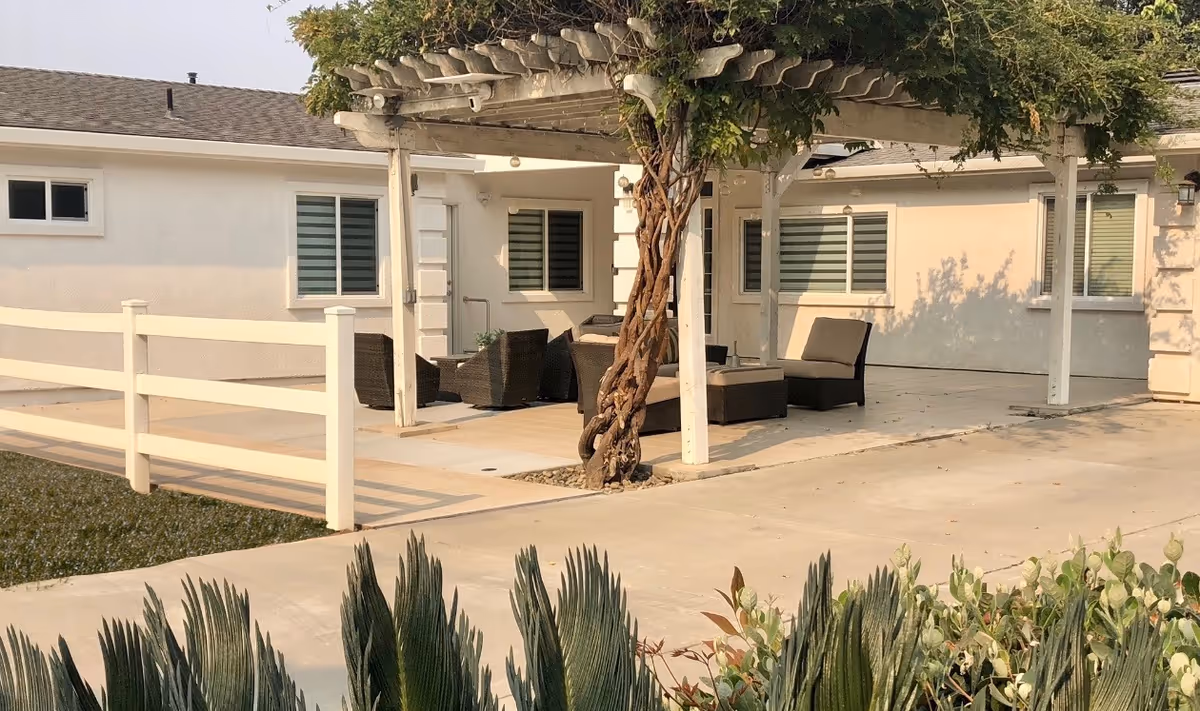 Outdoor patio area of a residential care facility with a pergola covered in greenery, outdoor wicker seating including chairs and a table, surrounded by a white fence and various plants in the foreground.