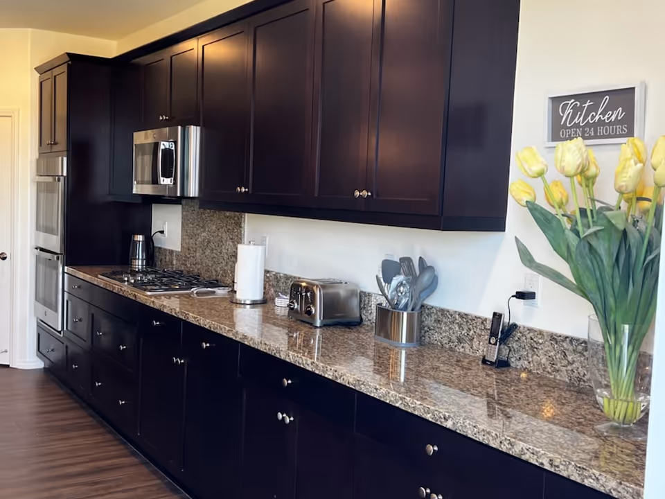 A modern kitchen with dark wood cabinets and granite countertops. The kitchen features a built-in microwave, oven, stovetop, toaster, paper towel holder, and a container with utensils. A vase with yellow tulips is placed on the right side of the counter under a sign that reads 'Kitchen OPEN 24 HOURS'.