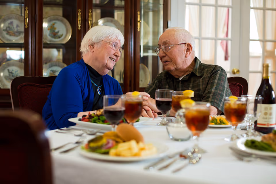 An elderly woman and man sitting at a dining table in a warmly lit room, holding hands and smiling at each other. The table is set with plates of food, glasses of iced tea with lemon, and a bottle of wine. Behind them is a wooden cabinet displaying decorative plates.