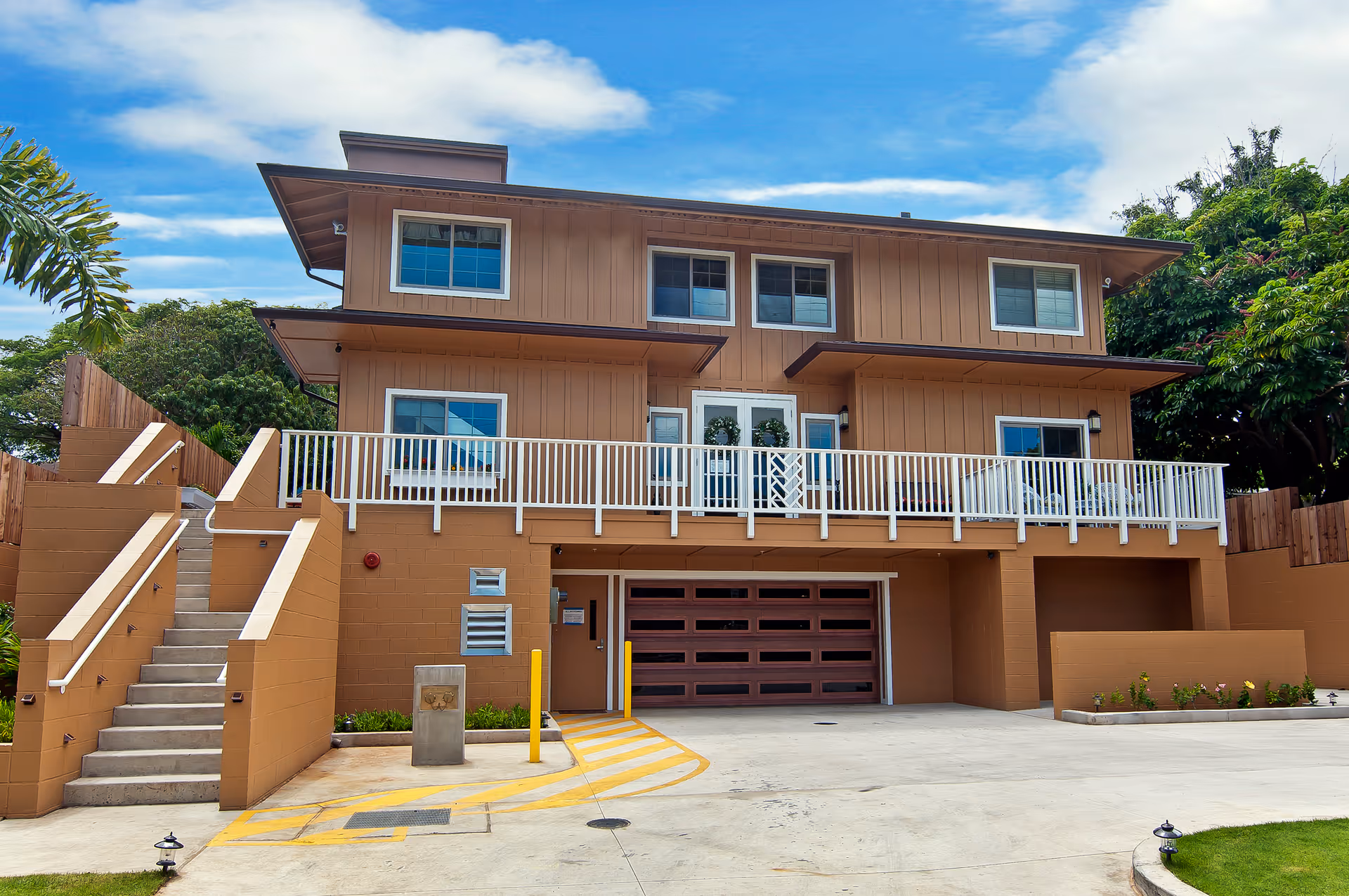 Exterior view of a two-story senior living facility building with a beige and brown color scheme. The building features multiple windows, a large garage door, a white railing balcony, and a staircase on the left side leading to the upper level. The surrounding area includes greenery and a clear blue sky with some clouds.