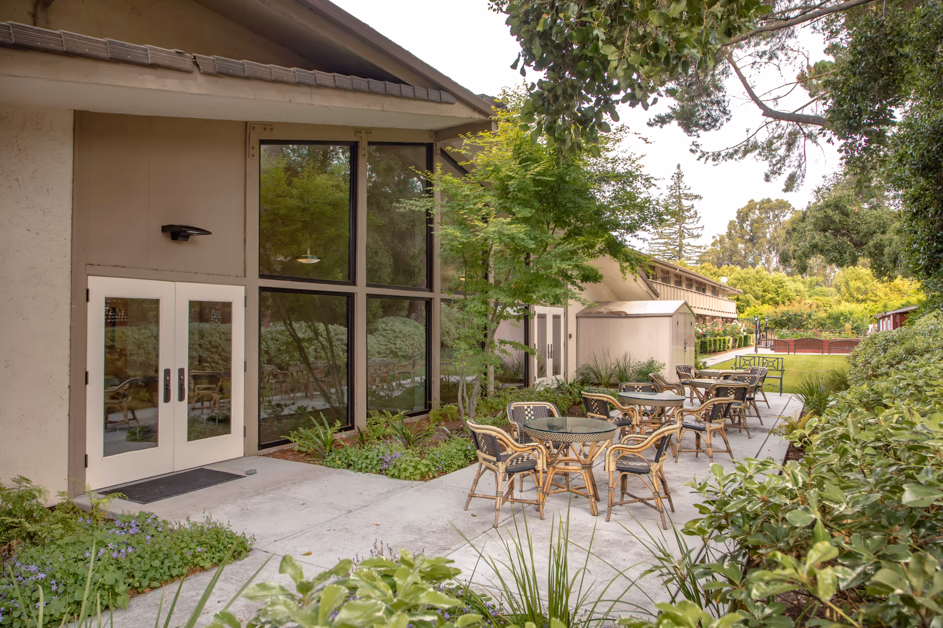 Outdoor patio area at BridgePoint at Los Altos with several round glass-top tables and wicker chairs arranged on a concrete walkway. The patio is adjacent to a building with large windows and double doors. Surrounding the patio are green shrubs, trees, and a well-maintained lawn extending into the background.