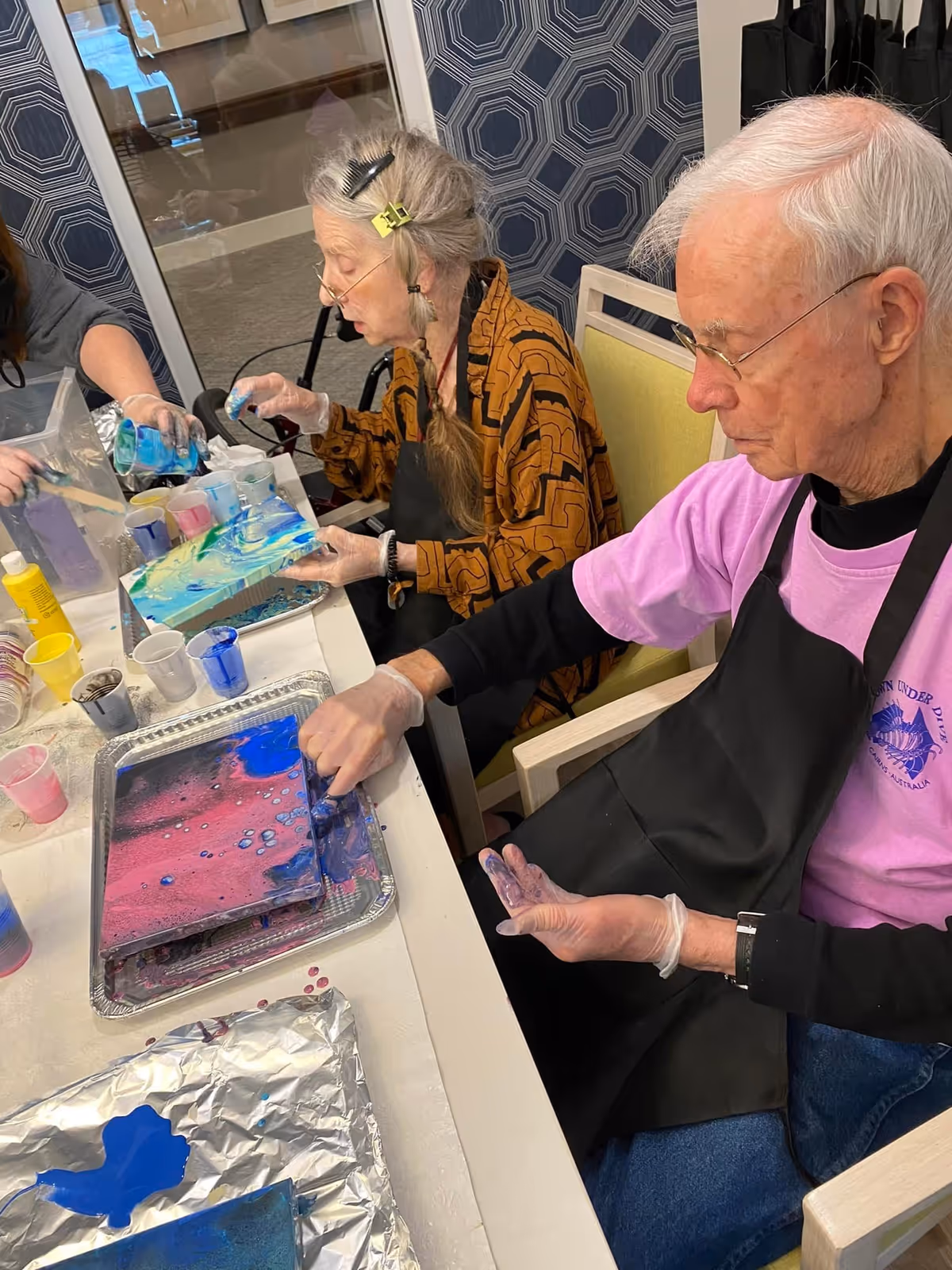 Two elderly individuals seated at a table engaged in a painting activity. They are wearing aprons and gloves, working with colorful paints on canvases. The setting appears to be indoors with patterned wallpaper in the background.