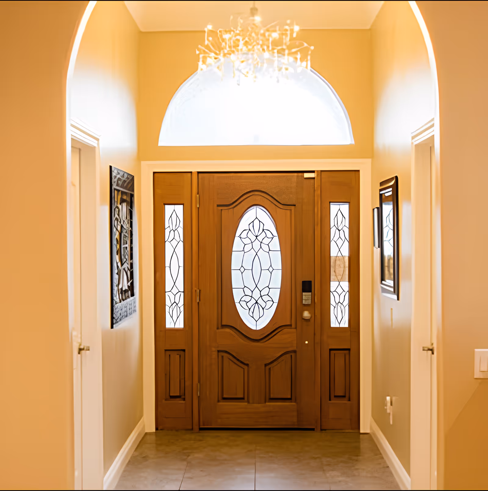Interior view of an entryway featuring a wooden front door with decorative frosted glass panels and sidelights. The walls are painted a warm beige color, and there is a chandelier hanging from the ceiling. Two doorways are visible on either side of the hallway, and framed artwork is hung on the walls.