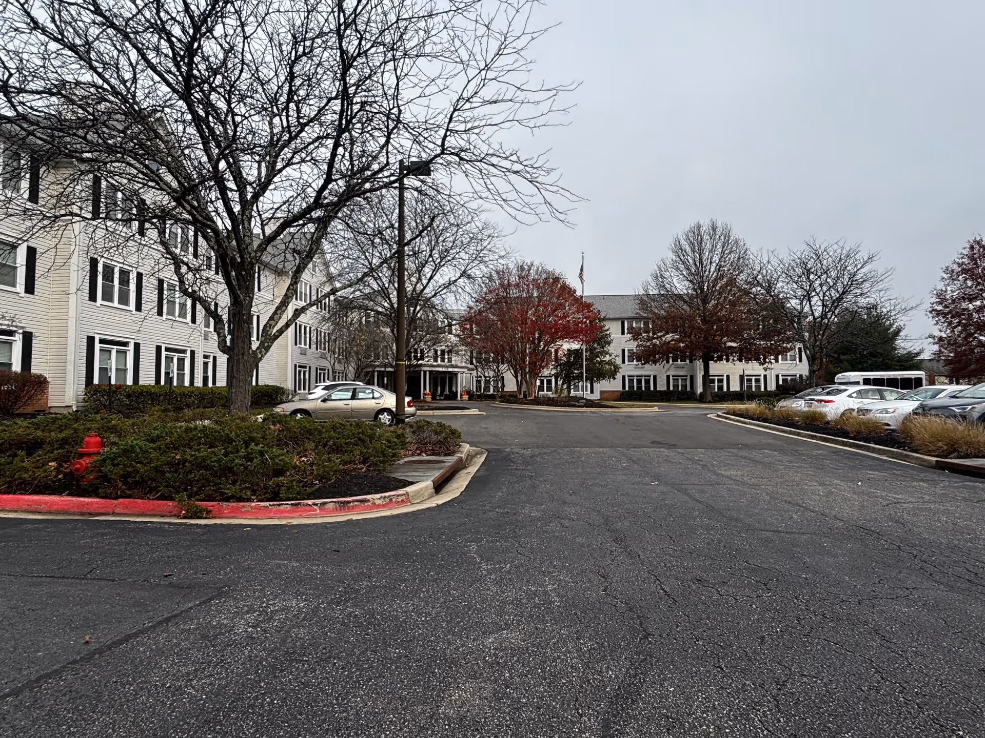 Exterior view of Morningstar Assisted Living facility showing a large parking lot with several parked cars, leafless trees, and a multi-story white building with black shutters under an overcast sky.