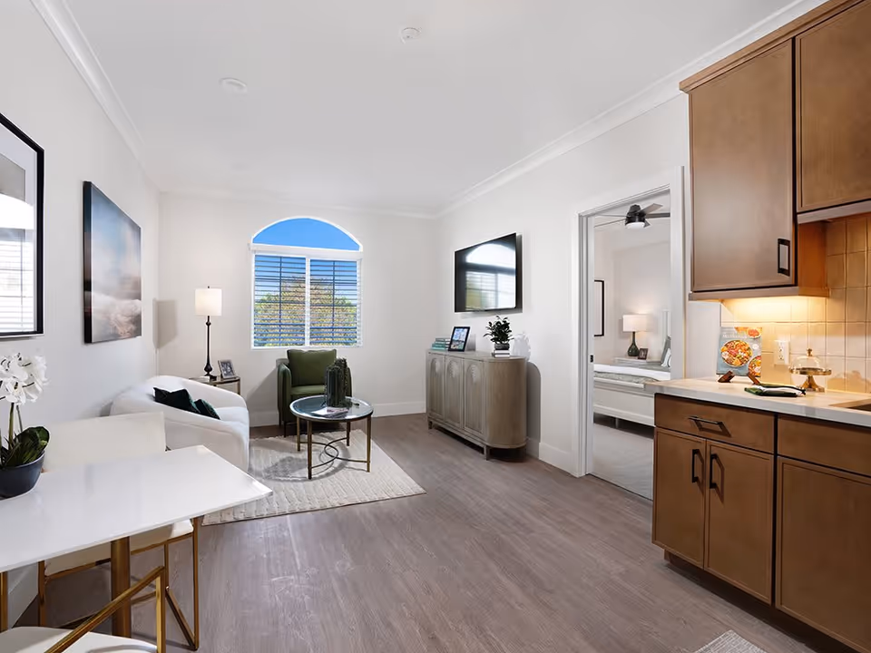 A bright living room with white walls and wood flooring. The room features a white sofa, a green armchair, a round glass coffee table on a light rug, a floor lamp, and a wall-mounted flat-screen TV above a wooden cabinet. A window with white shutters lets in natural light. To the right, there is a partial view of a kitchen area with wooden cabinets and a countertop. An open doorway leads to a bedroom with a bed, nightstand, and lamp visible.