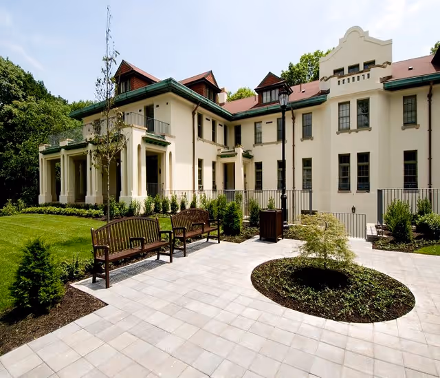 Outdoor patio area with two wooden benches, a small circular garden bed with a young tree, and a large cream-colored building with multiple windows and a red roof in the background, surrounded by greenery and trees.
