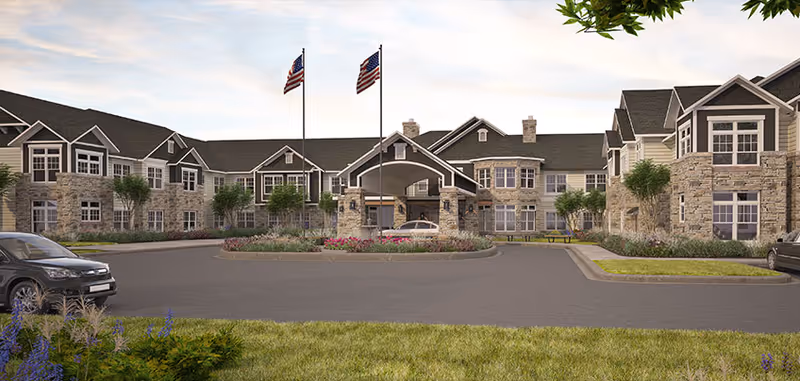 Front exterior view of a large senior living facility building with stone and siding facade, multiple windows, two American flags on flagpoles, a covered entrance with a circular driveway, and landscaped greenery and flowers.