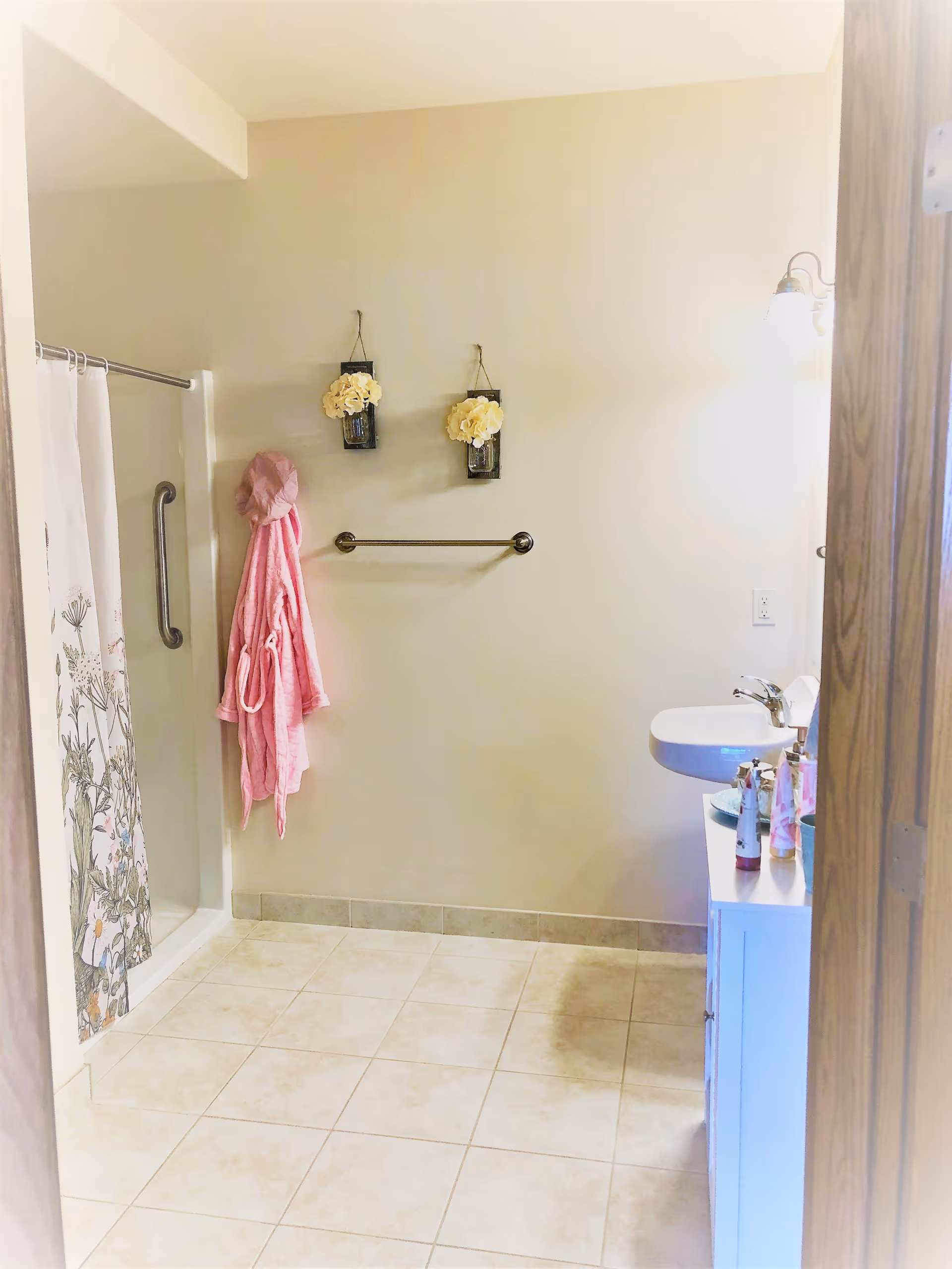 A bathroom with beige tiled floor and light beige walls. There is a shower with a floral curtain and a grab bar on the left side. A pink bathrobe is hanging on a hook next to the shower. Two small wall decorations with white flowers hang above a towel bar on the wall. On the right side, there is a white sink with a mirror above it and several bottles on the countertop. The bathroom is well-lit with a wall-mounted light fixture.