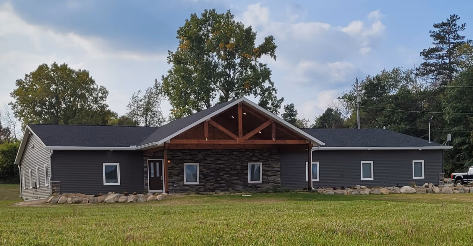 Exterior view of a single-story building with dark gray siding and a stone facade around the entrance. The entrance has a wooden gabled roof supported by wooden beams. The building is surrounded by grass and trees under a partly cloudy sky.